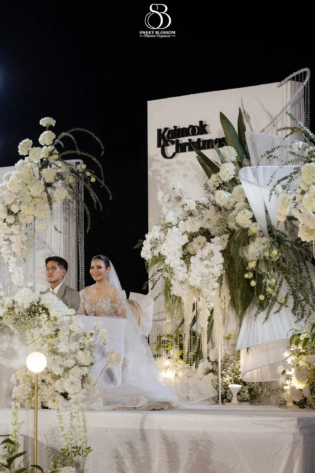 Couple seated at the white floral stage at the Mook — Christmas Wedding celebration in Lopburi