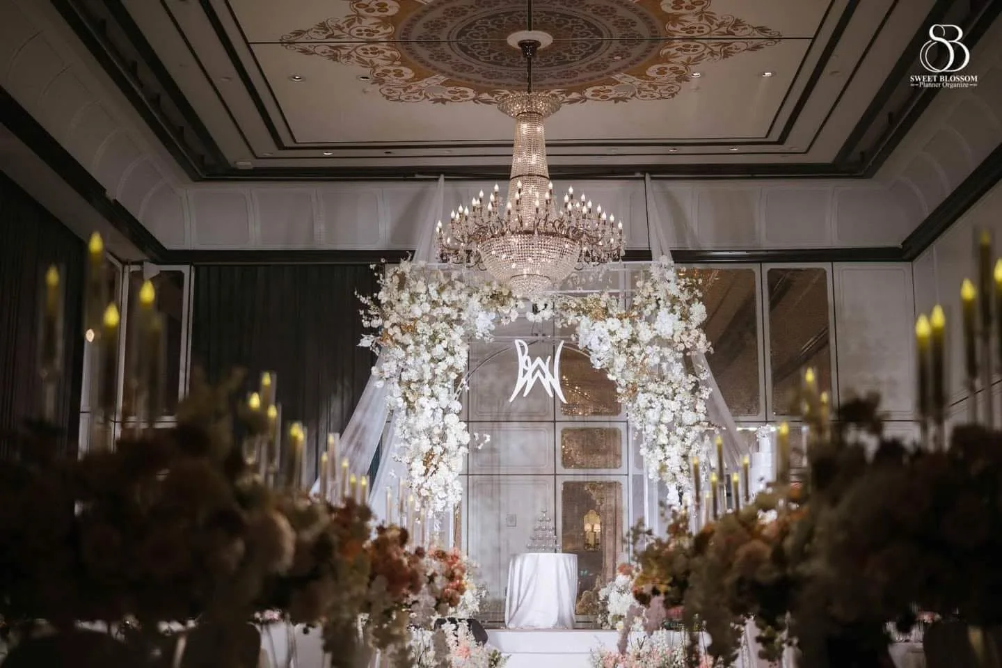 White floral ballroom stage beneath a crystal chandelier at the Risa & Ong celebration in Bangkok